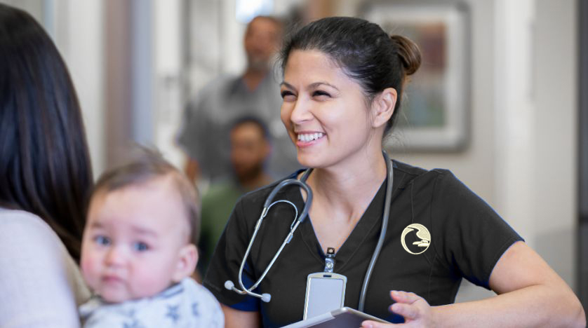 Family Nurse Practitioner smiling at a baby held by a parent in a clinic hallway.