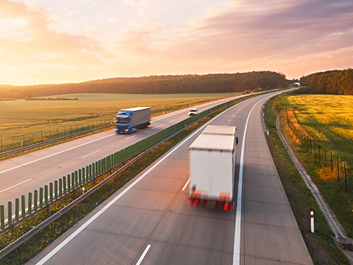 Trucks driving on a highway at sunset with open fields on either side, conveying movement and transportation.