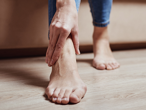 Close-up of a person's bare feet with one hand reaching down to touch the ankle, suggesting a focus on foot health or discomfort.