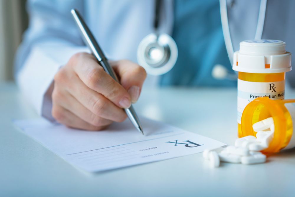 Close-up of a medical professional writing a prescription with a medication bottle and pills in the foreground.
